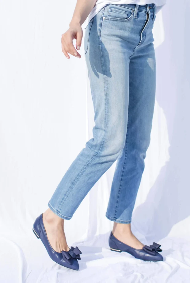 Close-up of legs in light blue jeans wearing navy bow-detail flats, on a plain white backdrop, calm and neutral mood